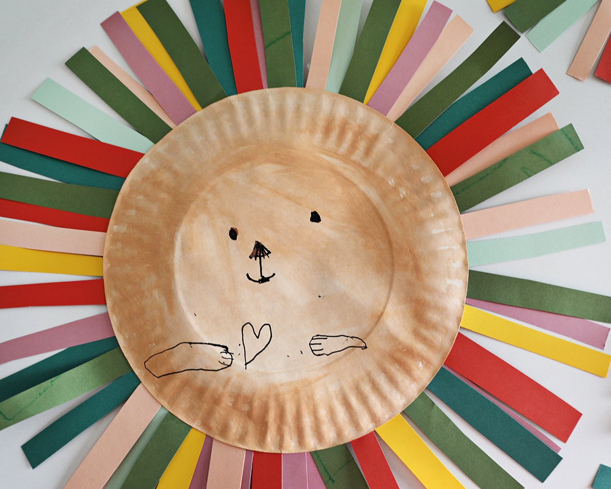 paper plate lion with rainbow strips of paper glued around it and a face drawn on. 