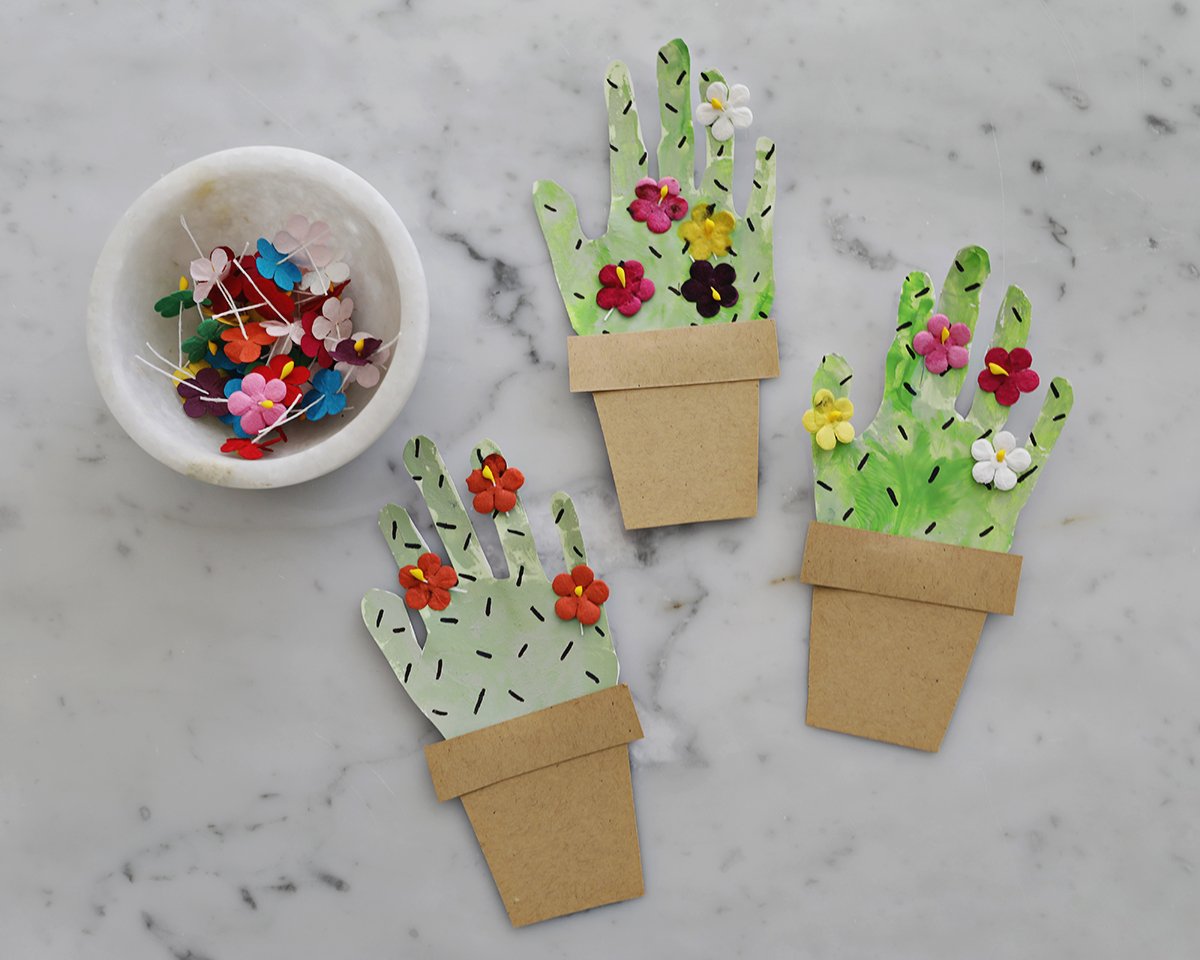 Cactus handprints in paper kraft paper pots on counter with bowl of craft flowers. 