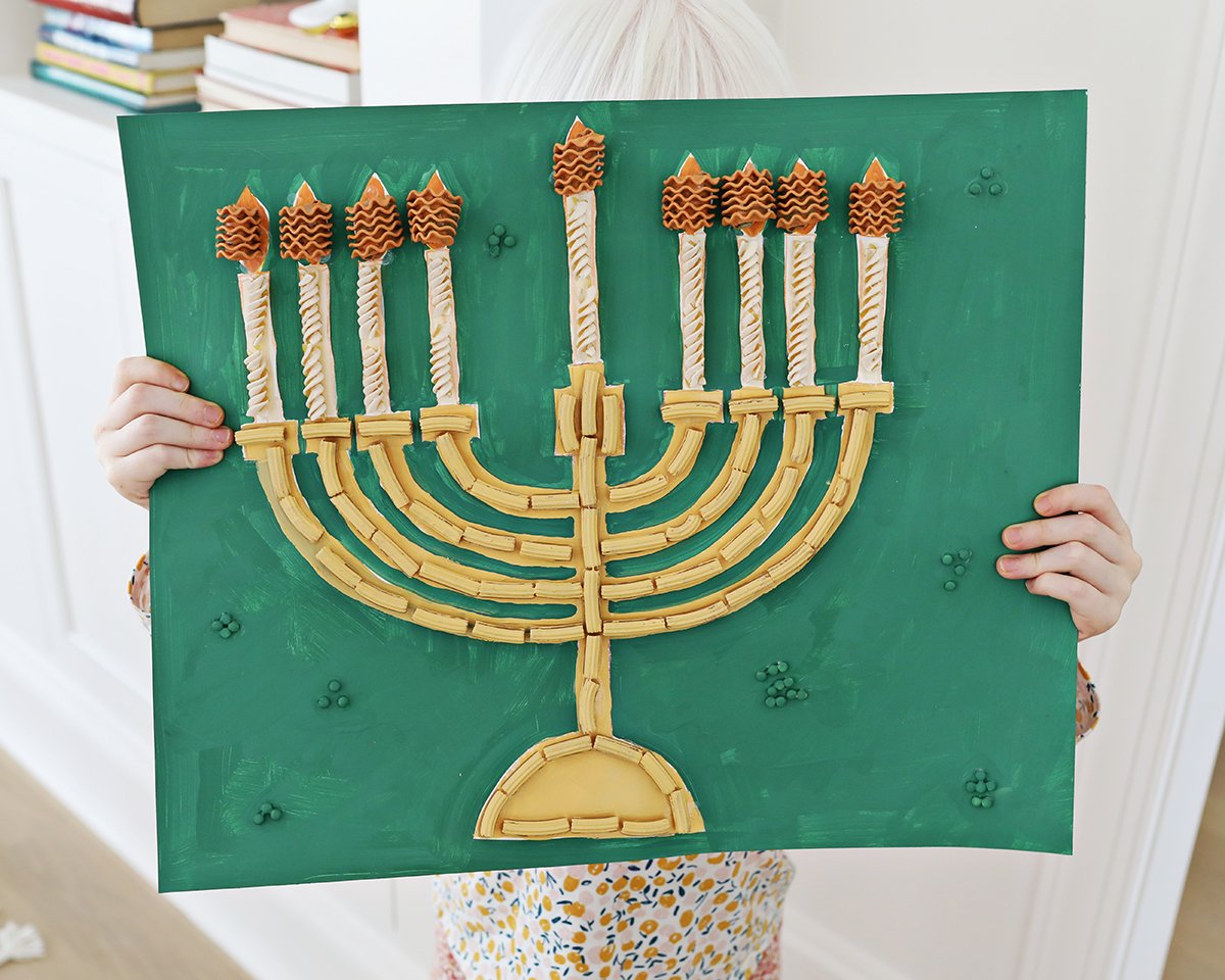 Child holding art project with noodles glued onto painted paper to make a menorah. 