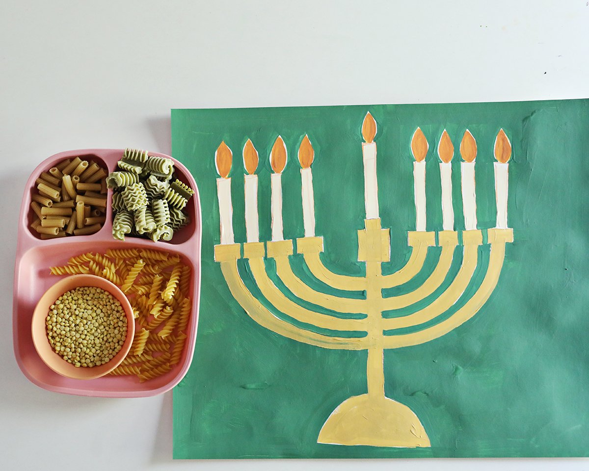 Painted menorah shape on green paper next to noodles in different shapes and sizes. 