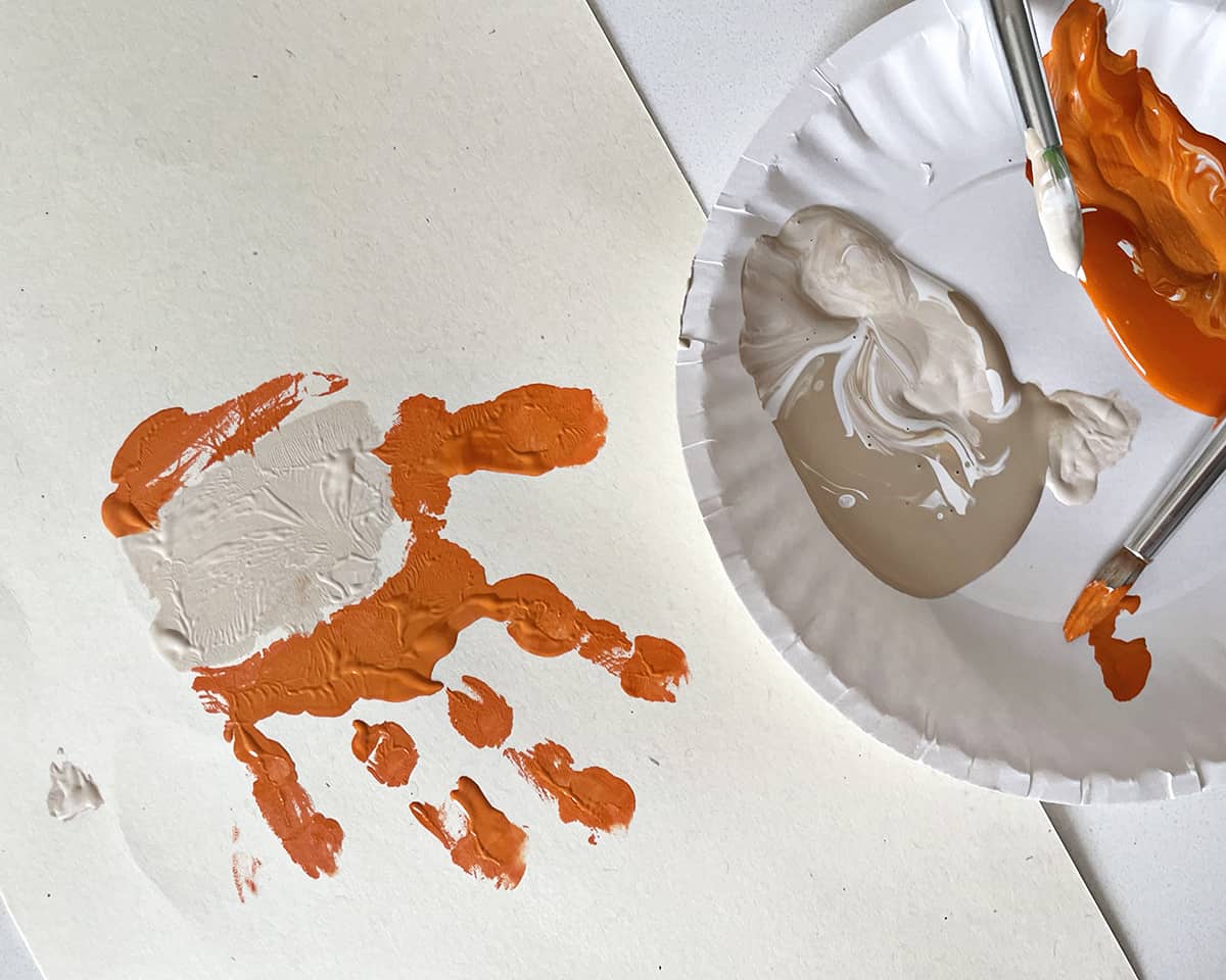 Orange and beige paint on plate and paintrbrush next to a child's handprint.