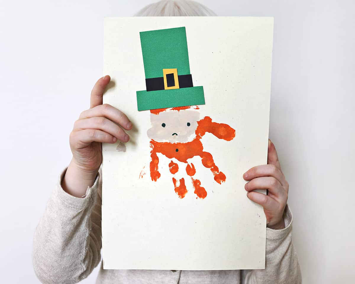 Child holding a crafted handprint leprechaun made with paint. 