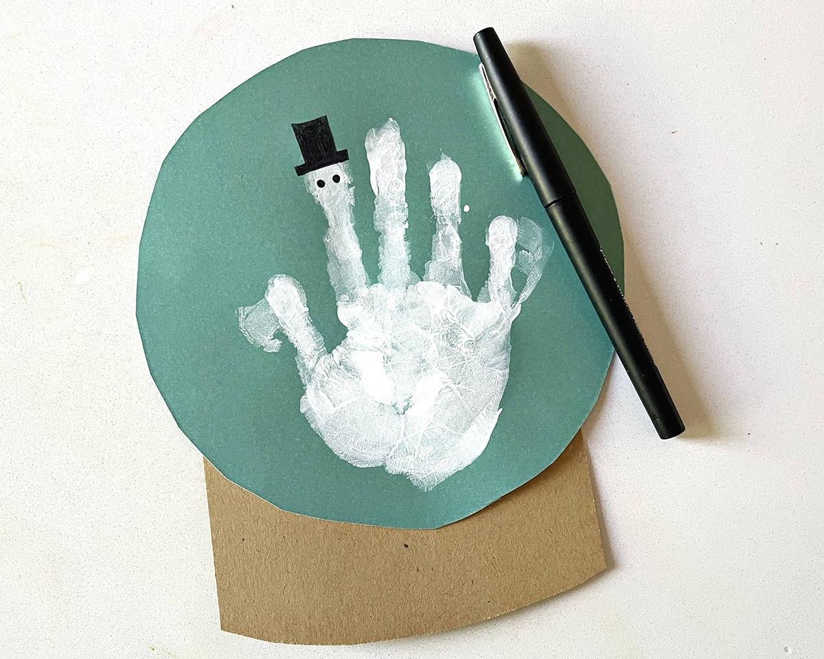 Snowman hat and face being drawn with a paint pen onto a white handprint on teal construction paper. 