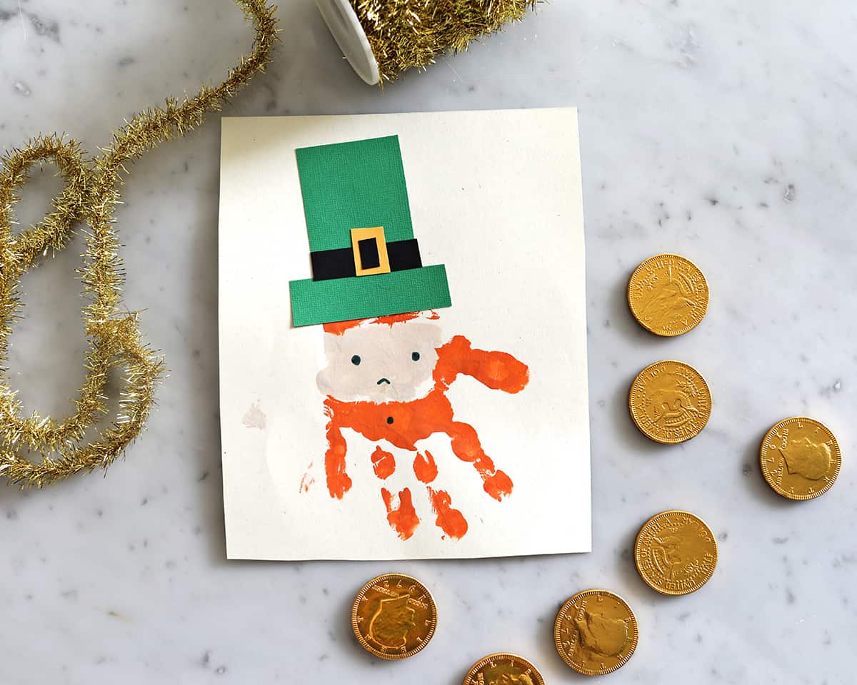 painted handprint on construction paper to look like a leprechaun next to gold coins on a counter. 