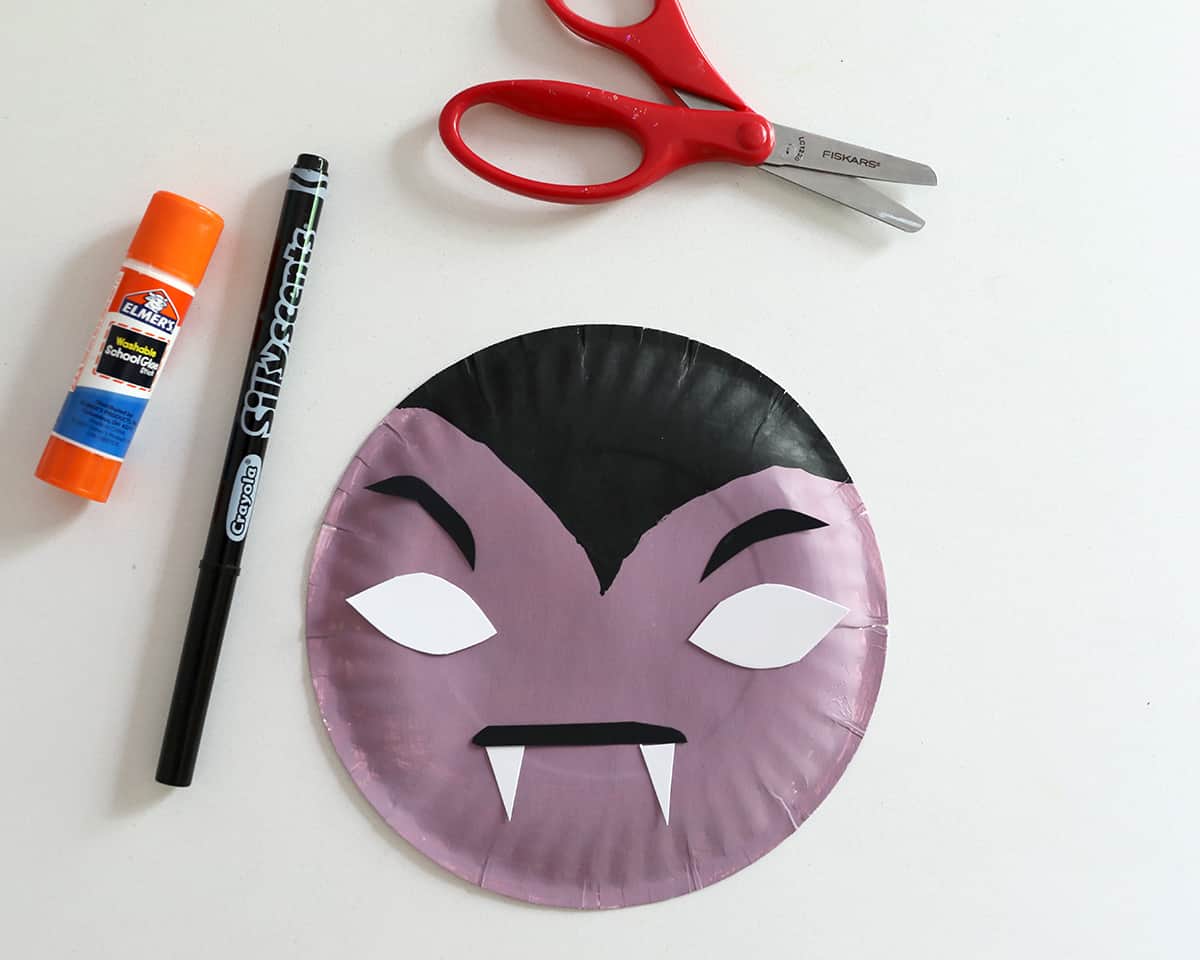paper plate painted purple and black next to glue stick, marker and scissors. 