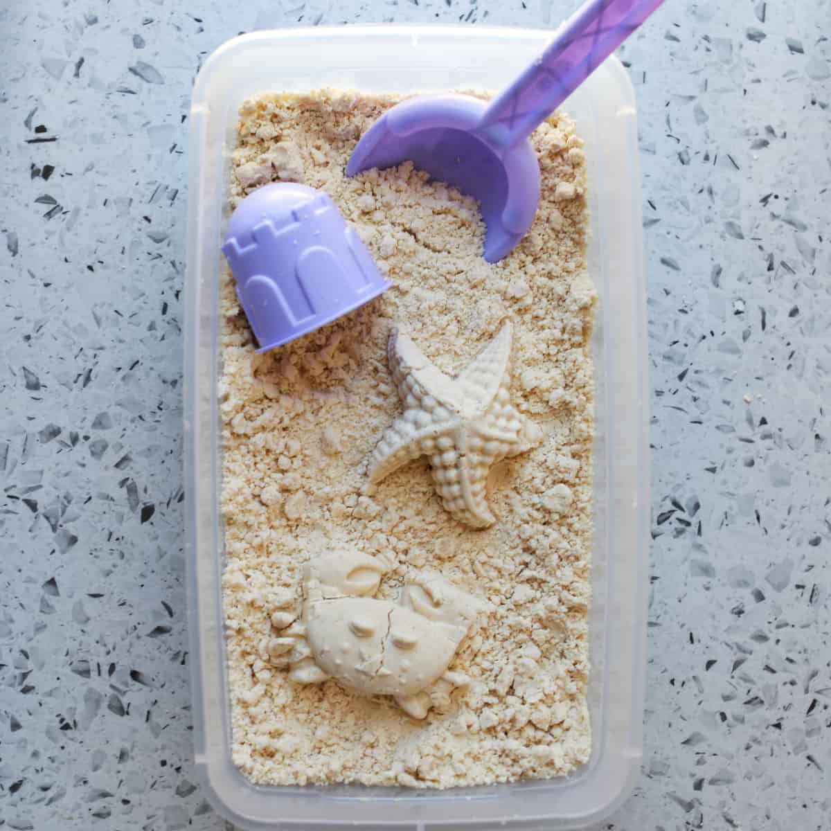Container holding moon sand with crab and starfish shape and purple scooper. 