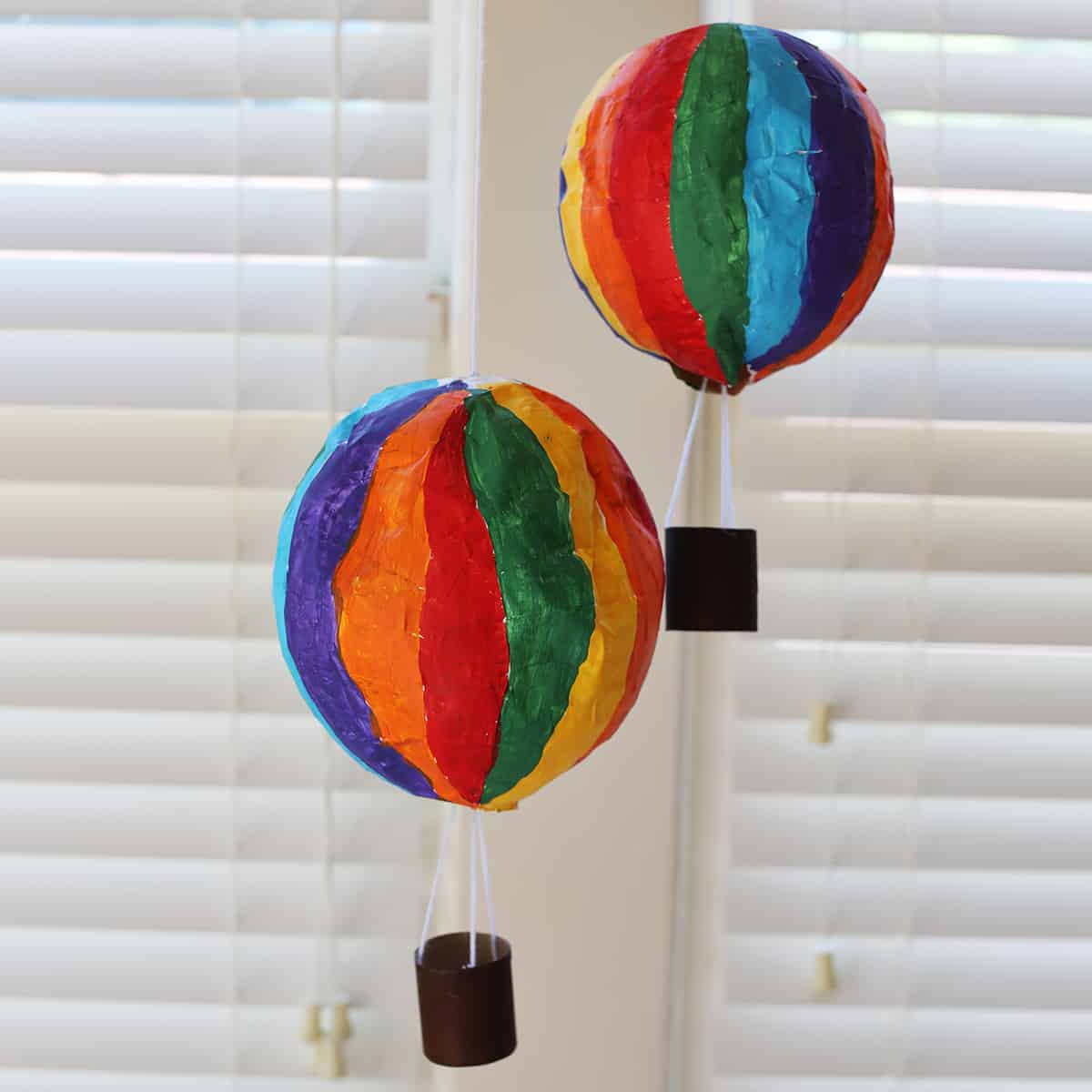 Hot air balloon craft painted with rainbow colors and tied with yarn to a painted brown paper towel roll. 