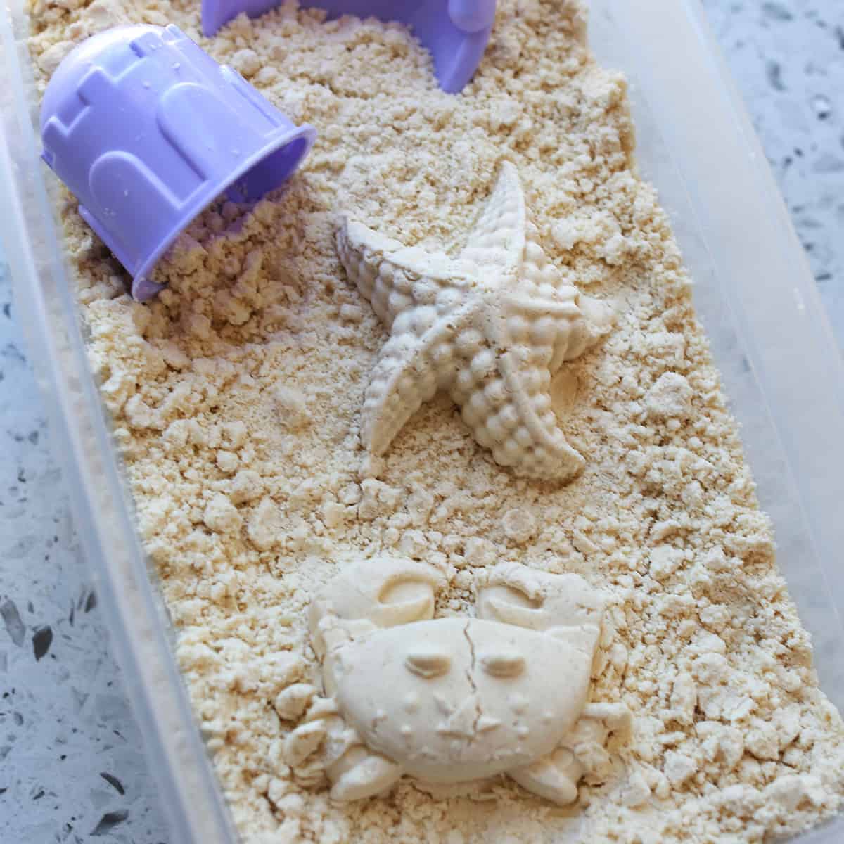 Moon sand shaped as starfish and crab in clear container with purple scoop. 