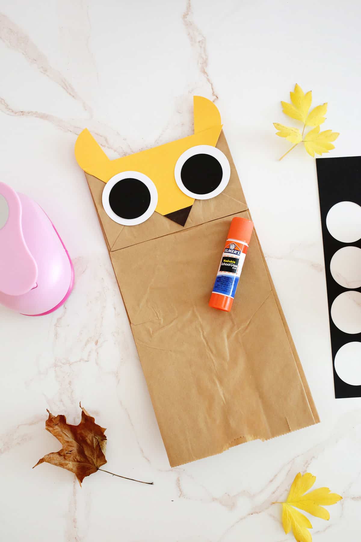 Glue stick sitting on brown paper bag with paper shapes to look like an owl. 
