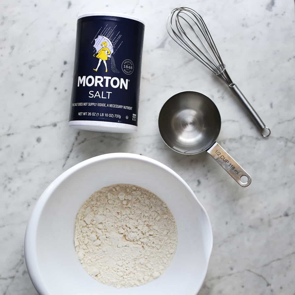 Whisk, measuring cup, salt and bowl with flour in it. 