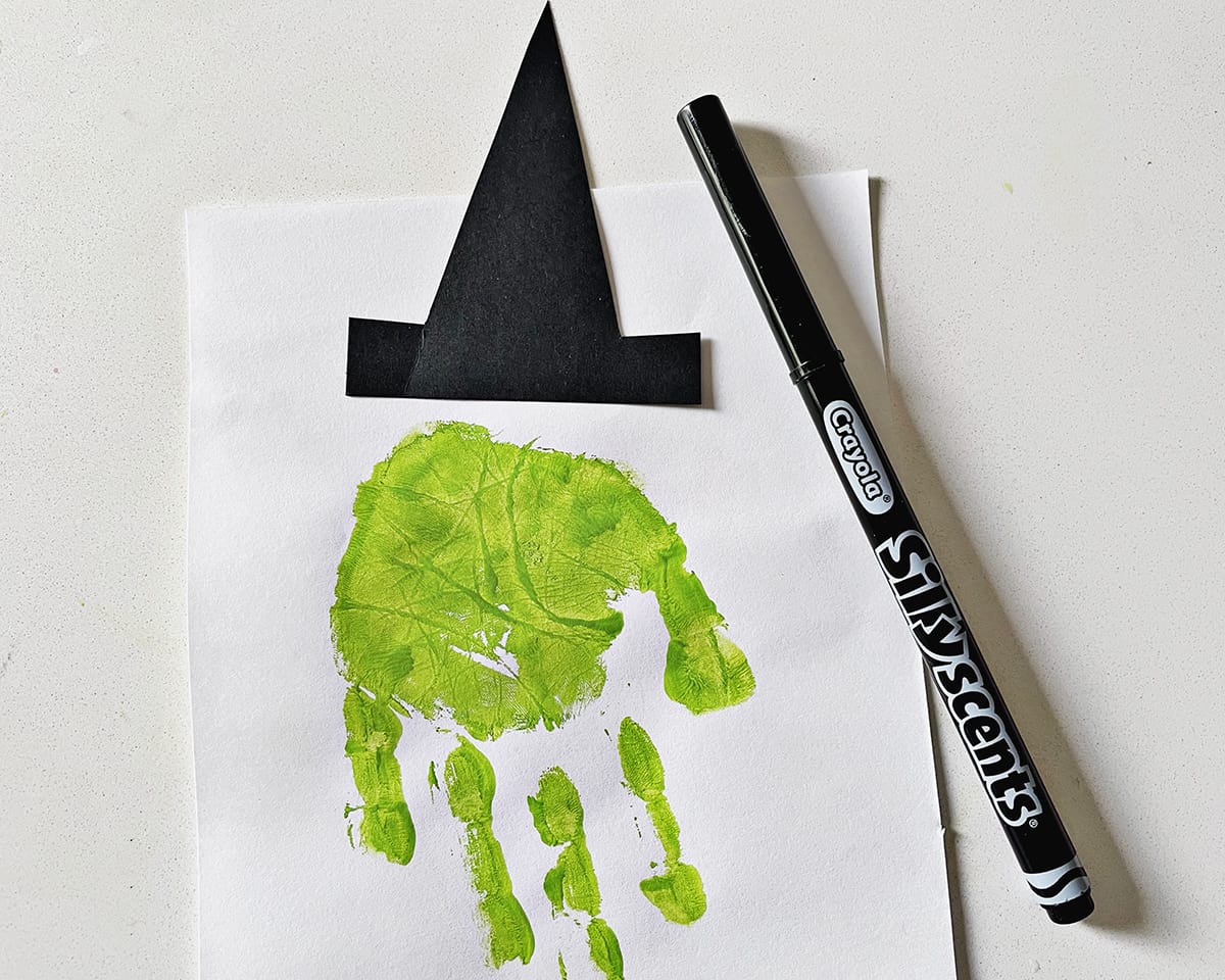 Black marker next to a piece of white construction paper with a green handprint and a black construction paper witch's hat. 