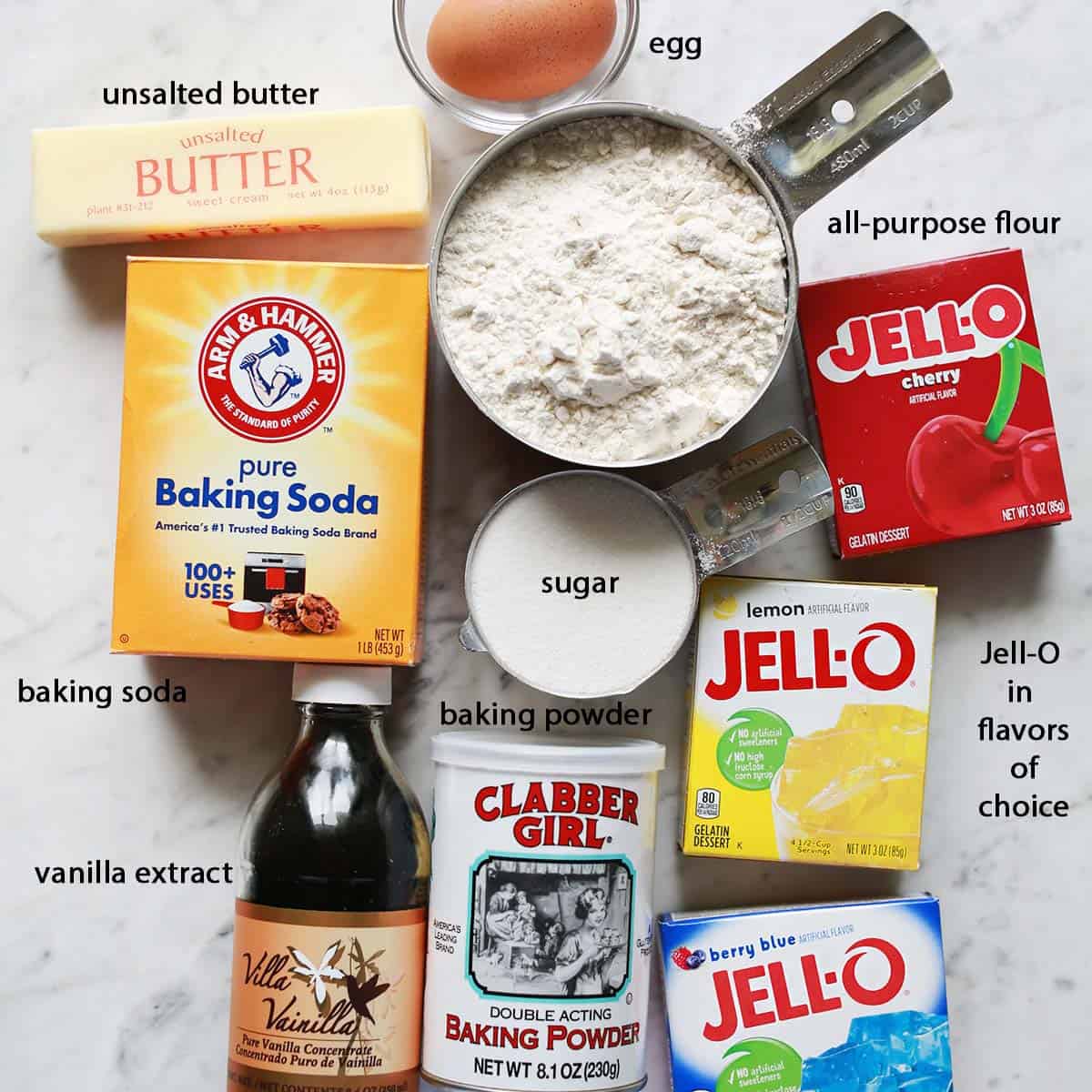 stick of butter, egg, cup of flour, box of baking soda, measuring cup of sugar, 3 boxes of jello, baking soda and a container of vanilla extract on a counter.