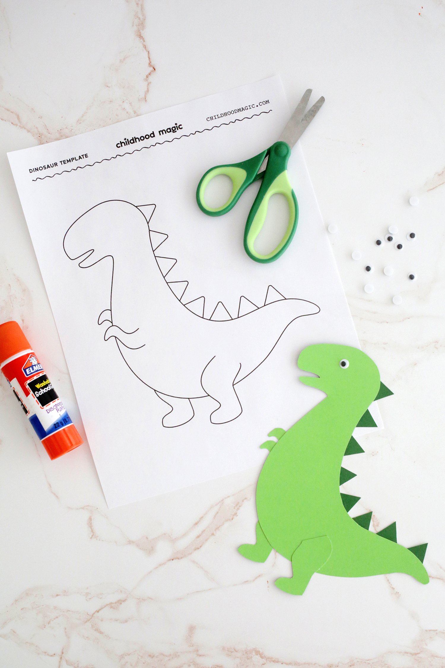 Green construction paper cut out in the shape of a t-rex and decorated next to a t-rex printable coloring page and a glue stick. 