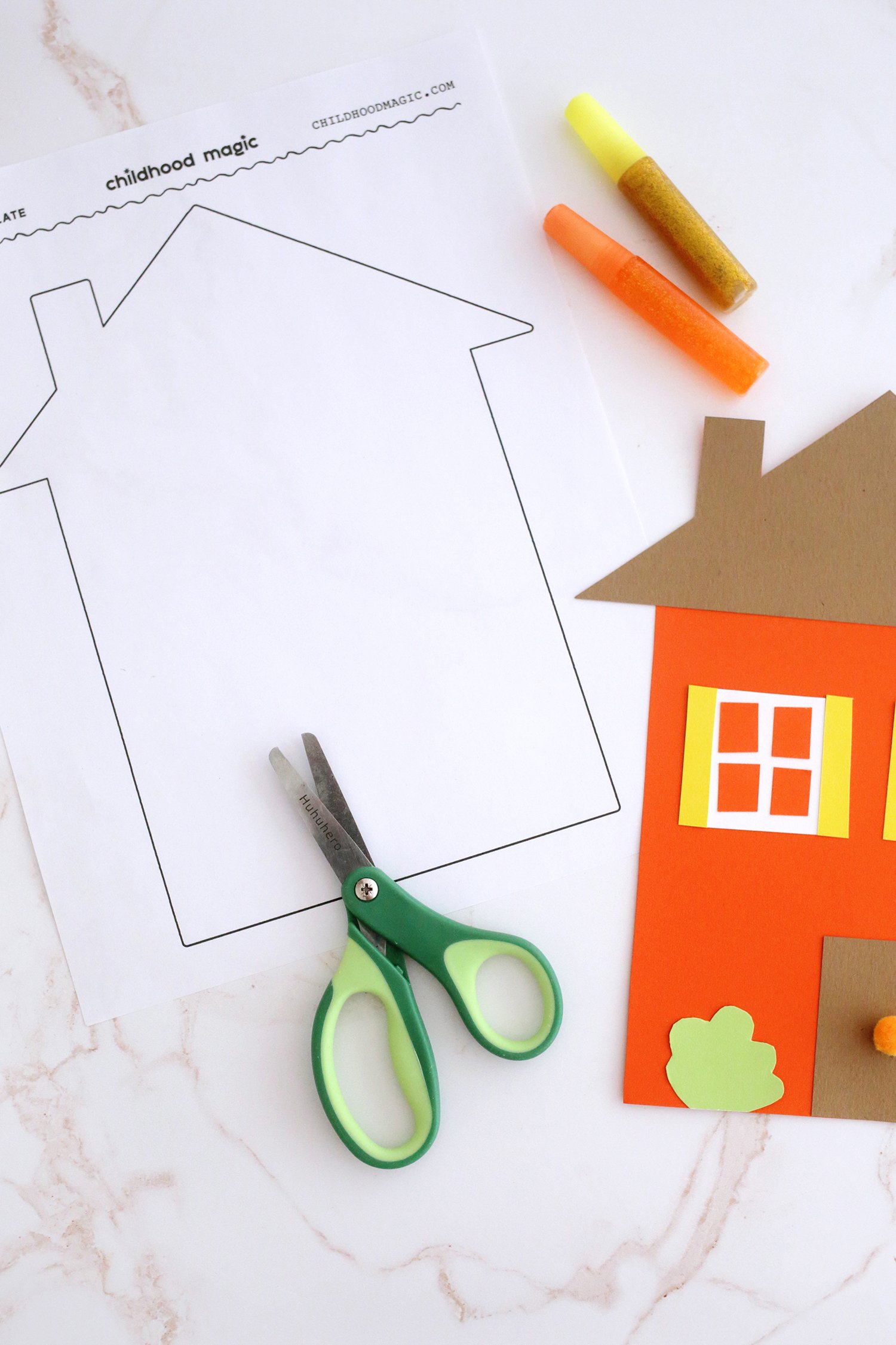 Green kids scissors next to an outline of a house on a white background. Glitter glue sticks and scraps of paper next to it. 