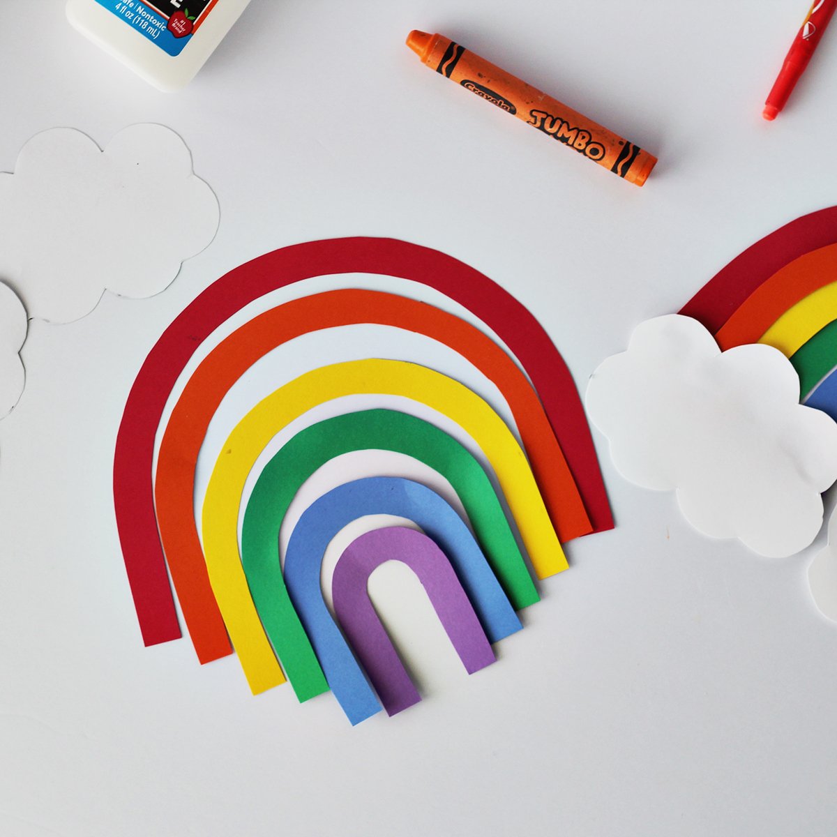 Red, orange, yellow, green, blue and purple strips of paper cut into arches to make a rainbow. The paper is next to an orange crayon, and white paper cut into cloud shapes.