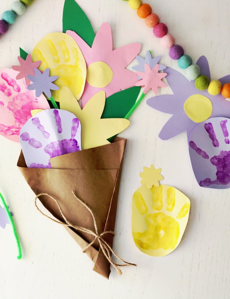 bouquet of flowers made from kids' handprints on pink, purple and yellow paper. Bouquet is wrapped in brown craft paper and sitting on a white table.