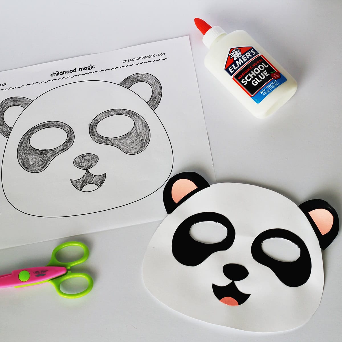 Mask made out of white, black and pink cardstock made to look like a panda. Next to the mask is a coloring page with the same mask on it. Next to the paper is a bottle of glue and a pair of scissors. 