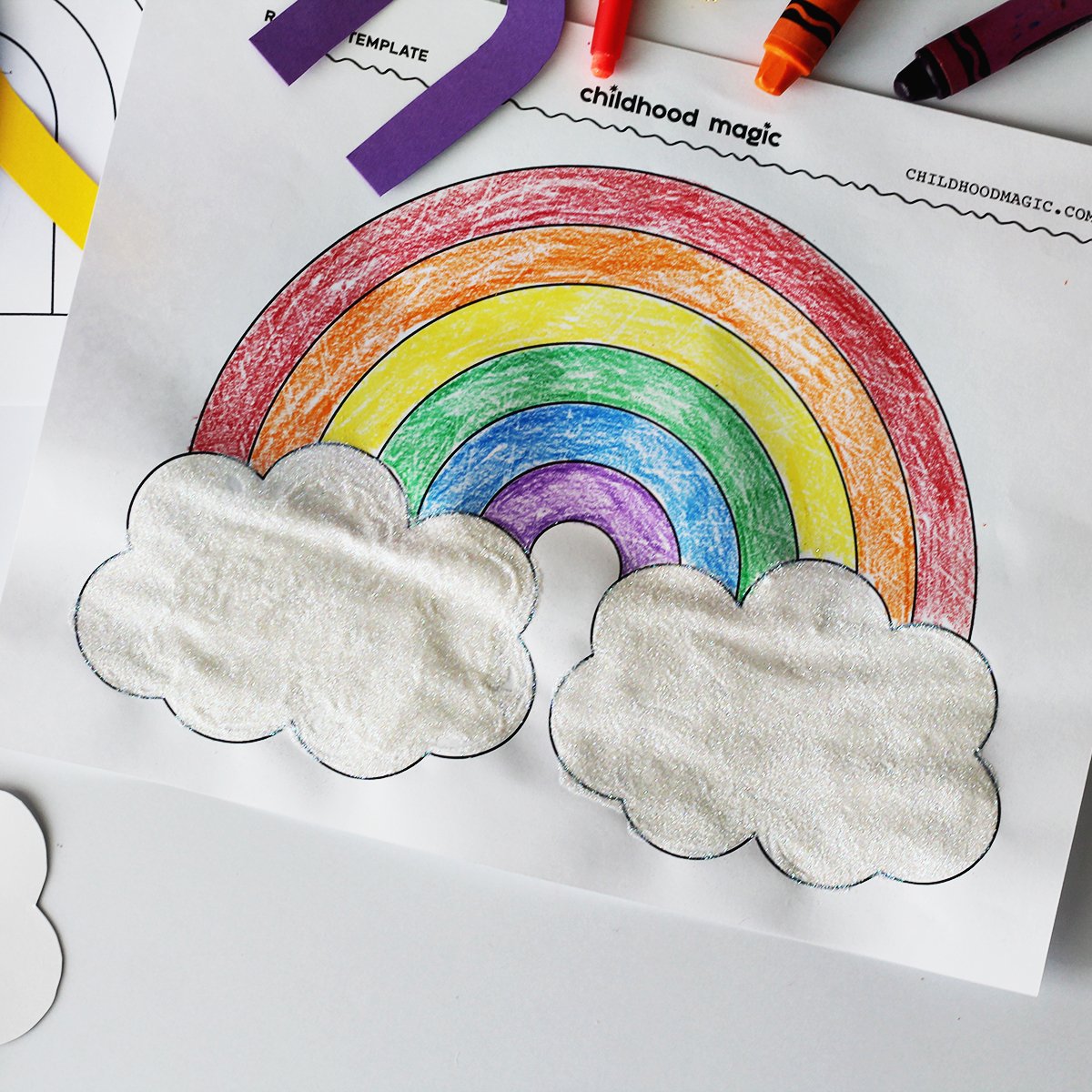 Coloring page with a rainbow shape colored in. Glitter on the page and crayons and purple paper next to the coloring page.