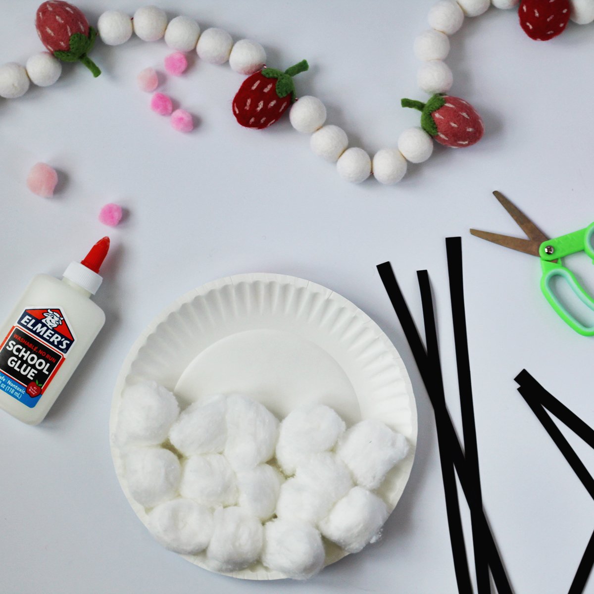 Poms being glued onto a paper plate next to black strips of paper, green kids' scissors and a bottle of kids glue.