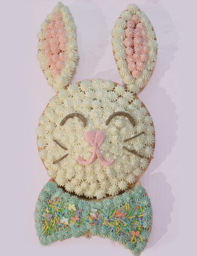 Cake in the shape of a rabbit with white, pink and blue frosting on a purple board.