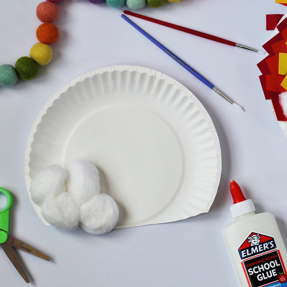 Paper plate with edge cut off and cotton balls glued to it. Next to the craft is a bottle of glue , red and blue paint brushes and green kids scissors.