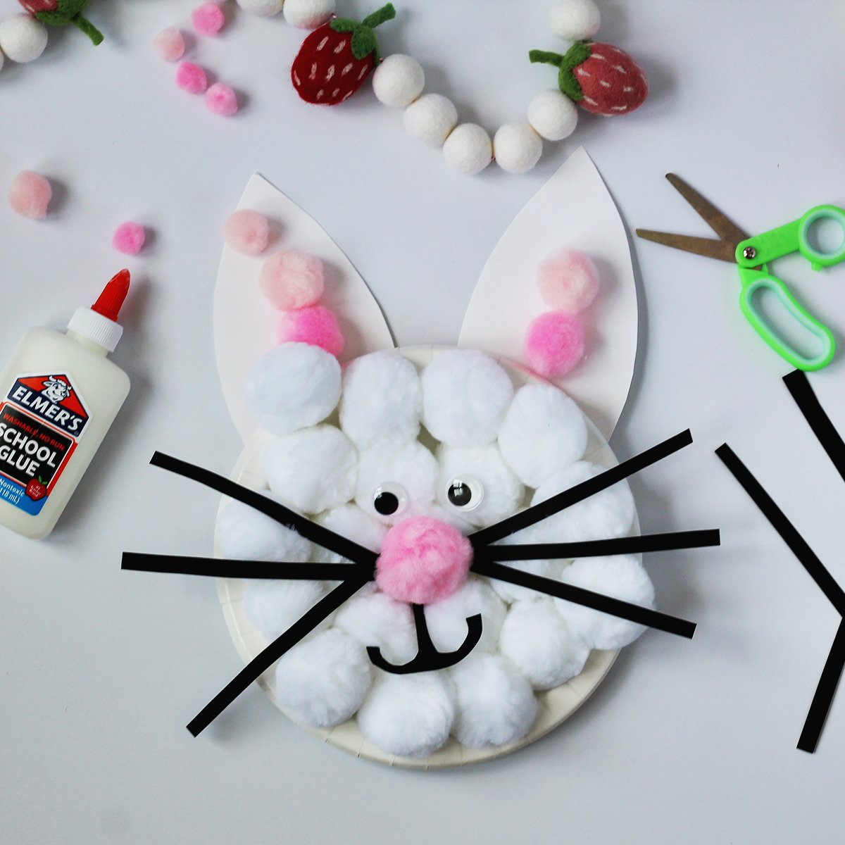 White plate with poms glued to it to make a rabbit face. Next to the bunny is a bottle of school glue and green kids' scissors.