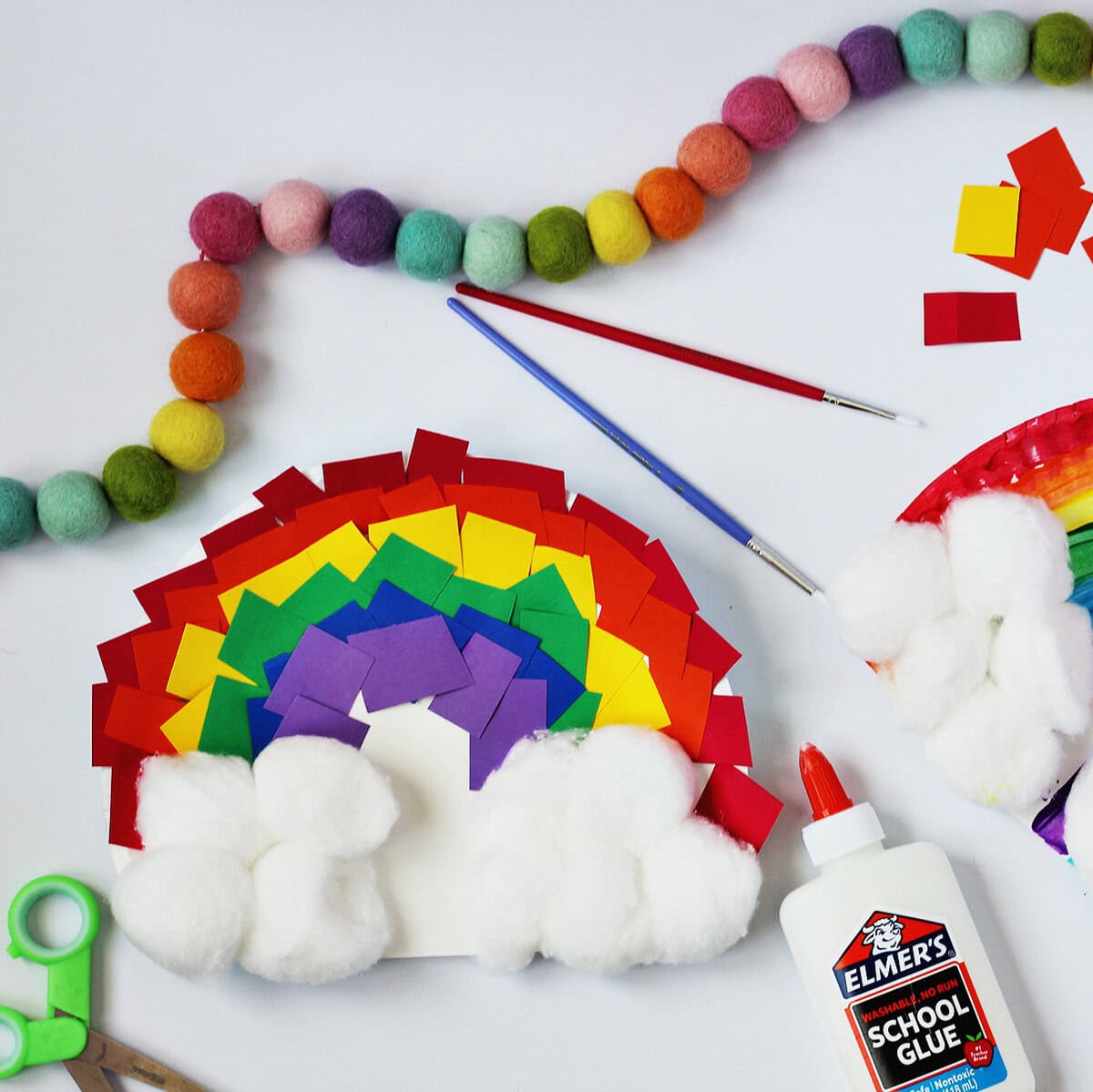 Rainbow craft made from a paper plate and scraps of paper in red, orange, yellow, green, blue and purple. Next to the craft is a bottle of glue , red and blue paint brushes and green kids scissors.