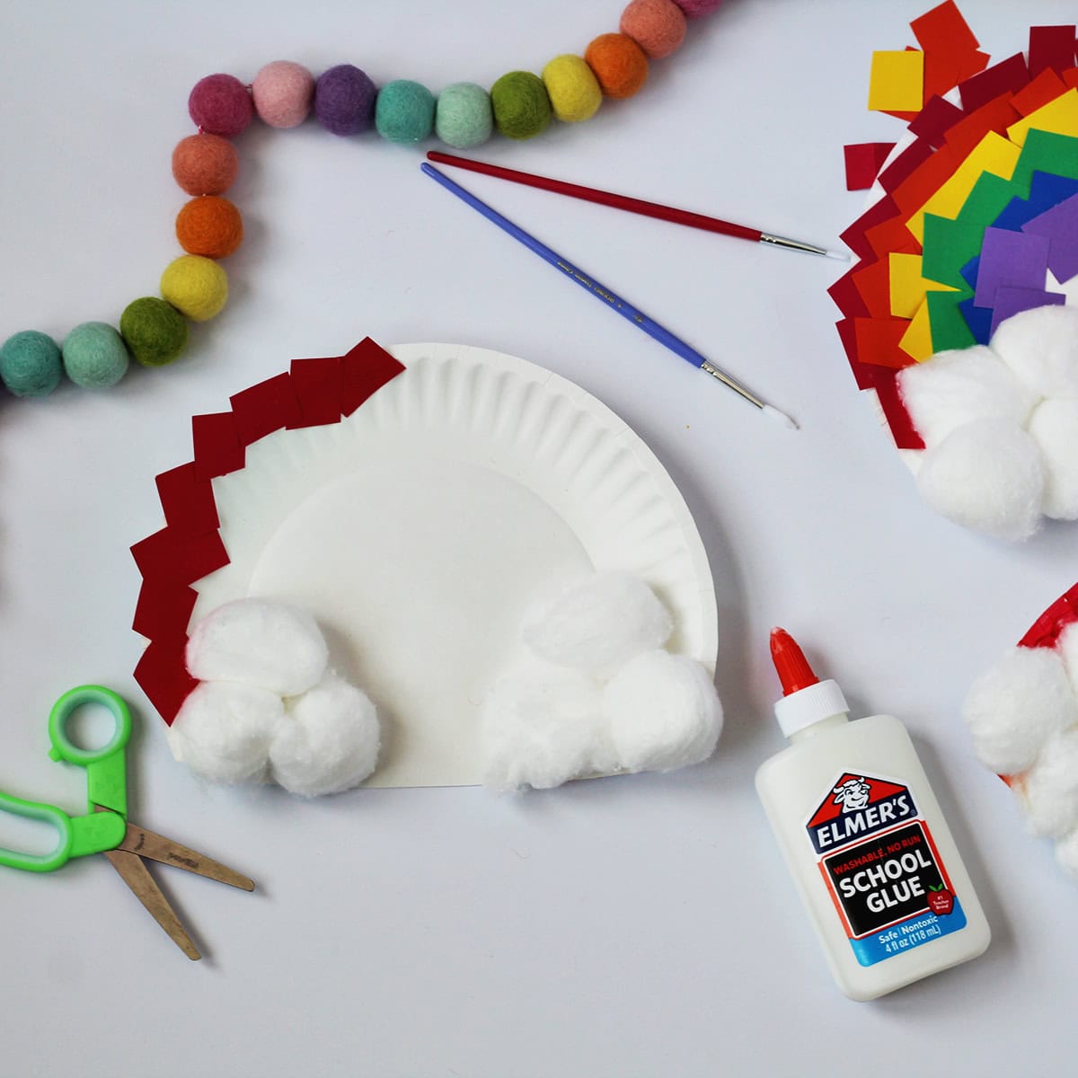 Paper plate with cotton balls glued onto the bottom and red paper glued around the top edge. Next to the plate is a bottle of glue and a pair of green kids scissors.