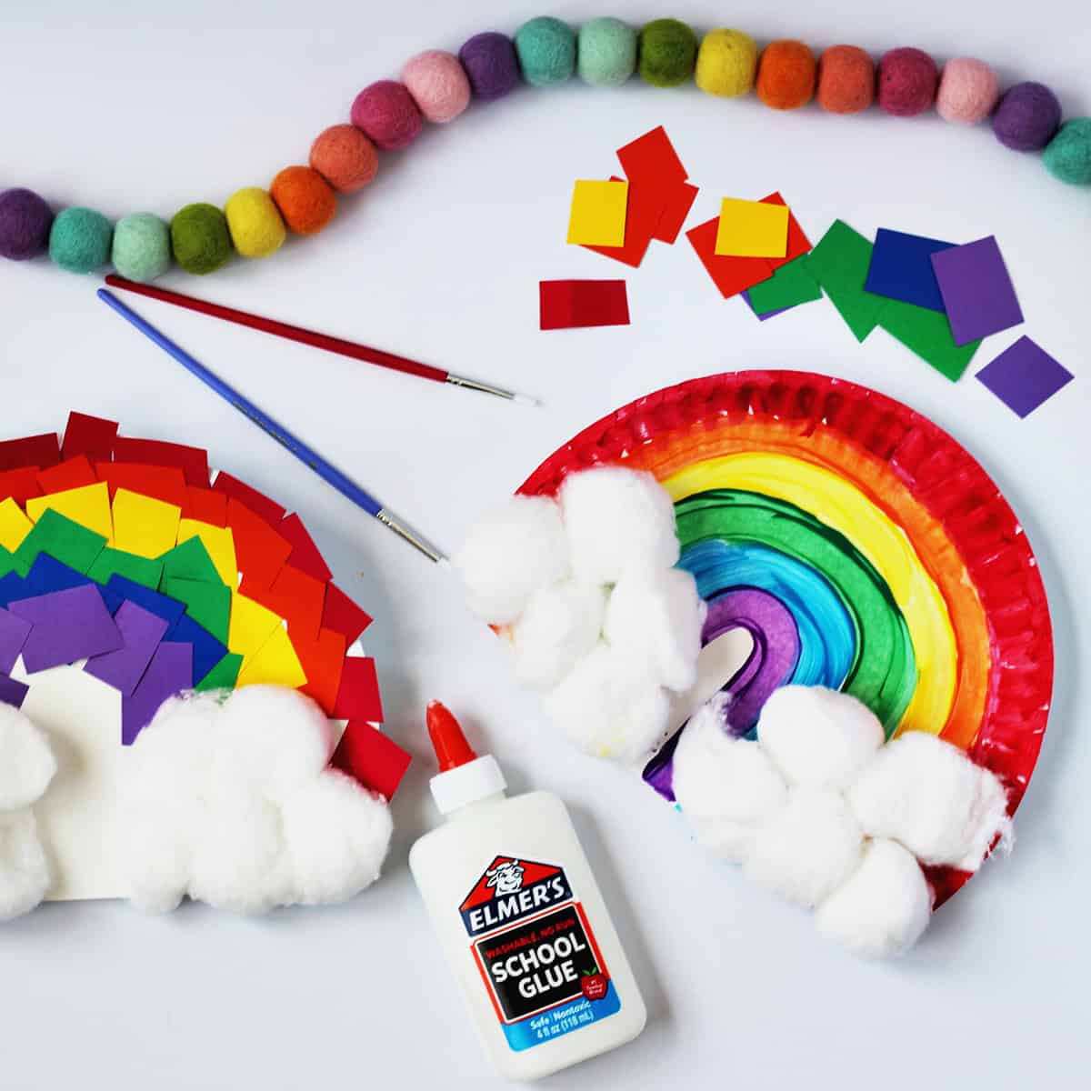 Rainbow craft made from a paper plate and scraps of paper in red, orange, yellow, green, blue and purple. Next to the craft is a bottle of glue , red and blue paint brushes and green kids scissors. Another paper plate rainbow is shown with painted stripes on it and cotton balls glued to the bottom.