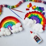 Rainbow craft made from a paper plate and scraps of paper in red, orange, yellow, green, blue and purple. Next to the craft is a bottle of glue , red and blue paint brushes and green kids scissors. Another paper plate rainbow is shown with painted stripes on it and cotton balls glued to the bottom.