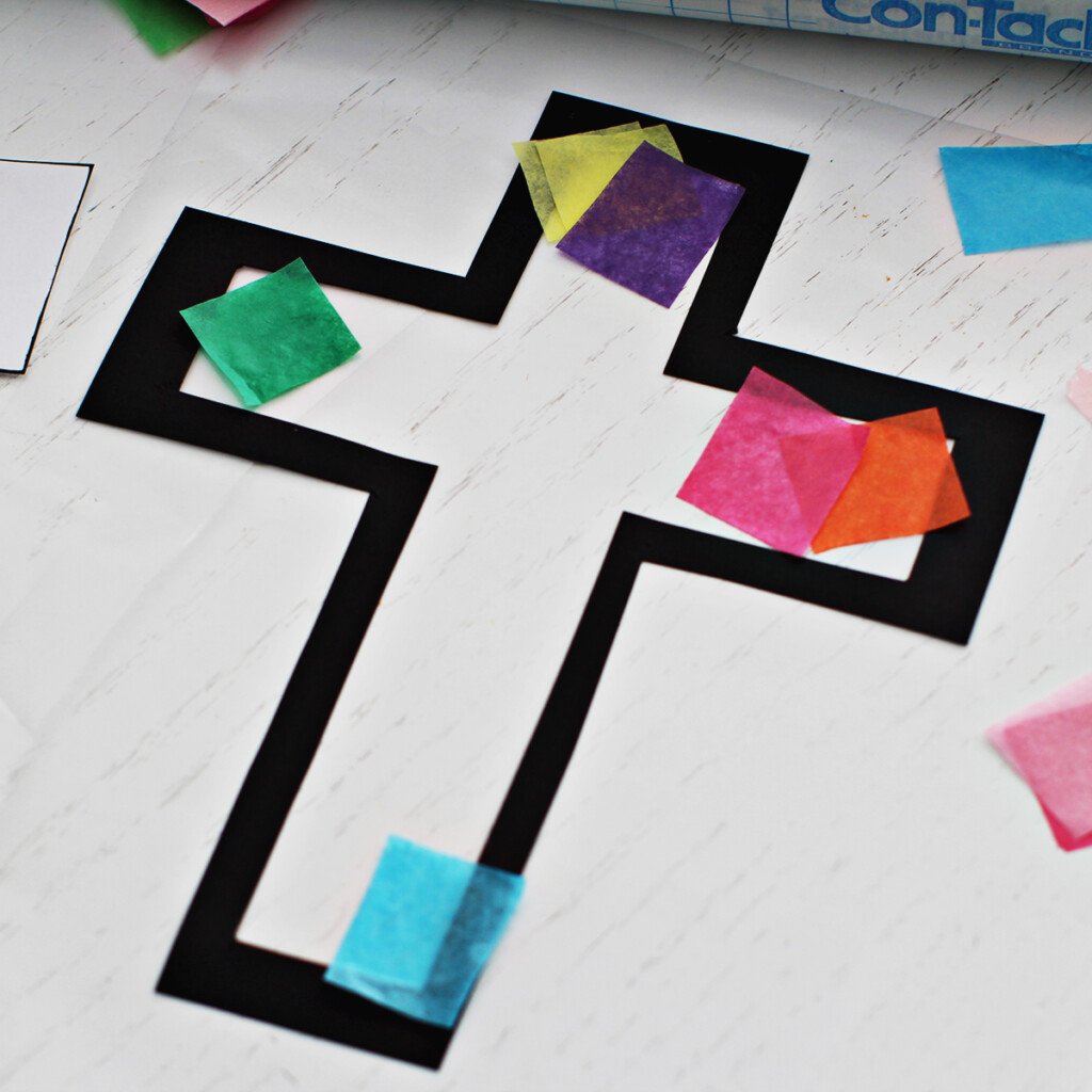 Colored tissue paper being added to a cross outline on contact paper to make a suncatcher or stained glass cross craft.