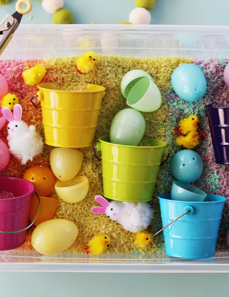 Plastic bin filled with rice dyed in rainbow pastel colors. Plastic eggs and toy chicks and bunnies are also in the plastic bin.