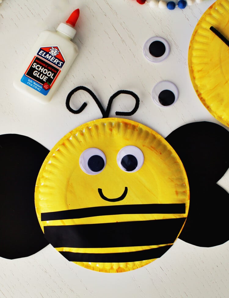 Paper plate painted yellow with black cardstock and googley eyes glued onto it to make a bumblee craft. A bottle of school glue is sitting next to the craft.