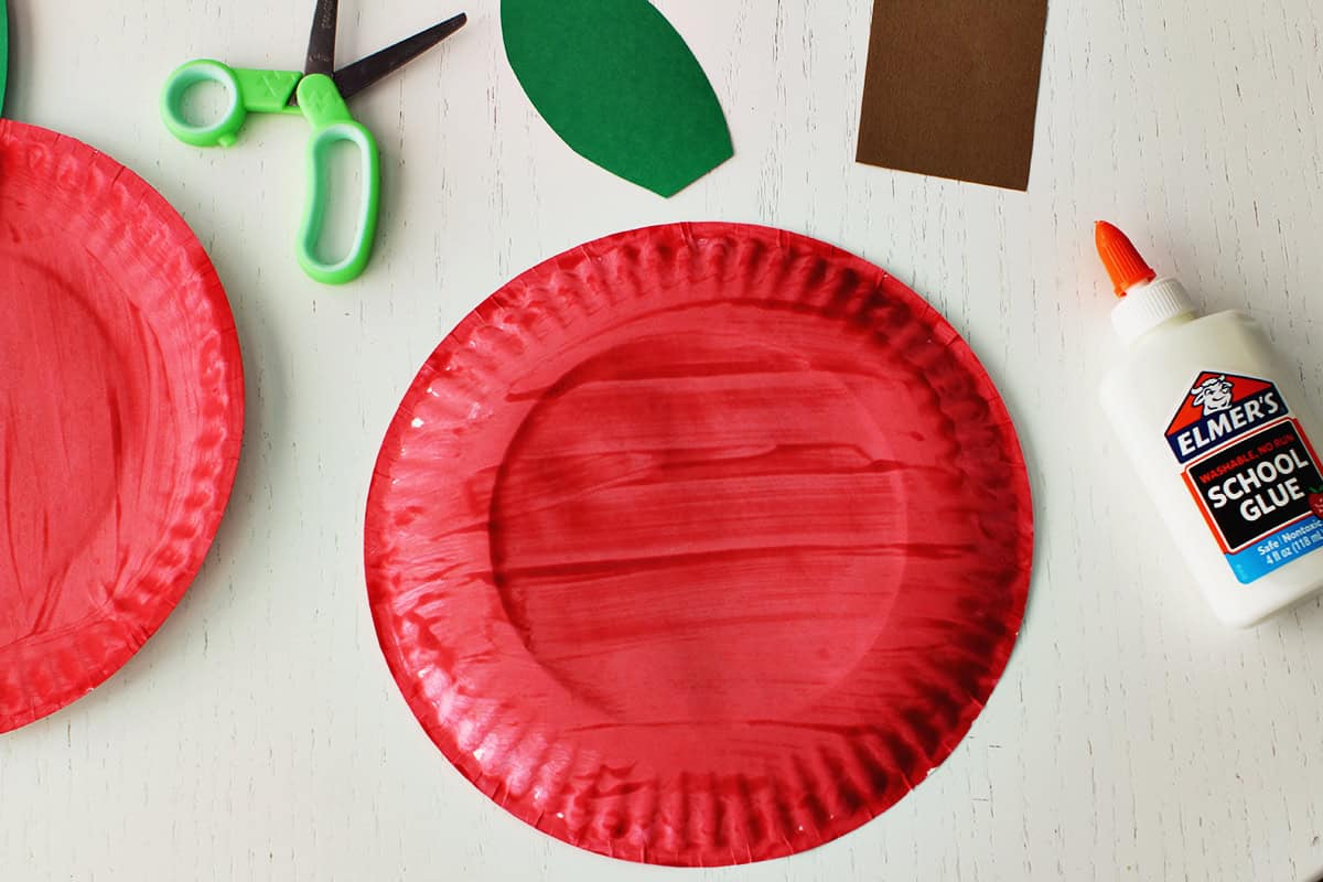 Red painted plate next to a bottle of school glue and a pair of green scissors. Also on the table is a piece of brown cardstock and a green cardstock cut into a leaf shape.