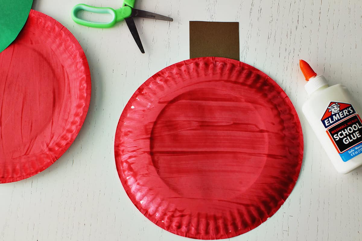 Red painted plate with a brown cardstock stem next to a pair of green kids scissors and a bottle of Elmer's glue.