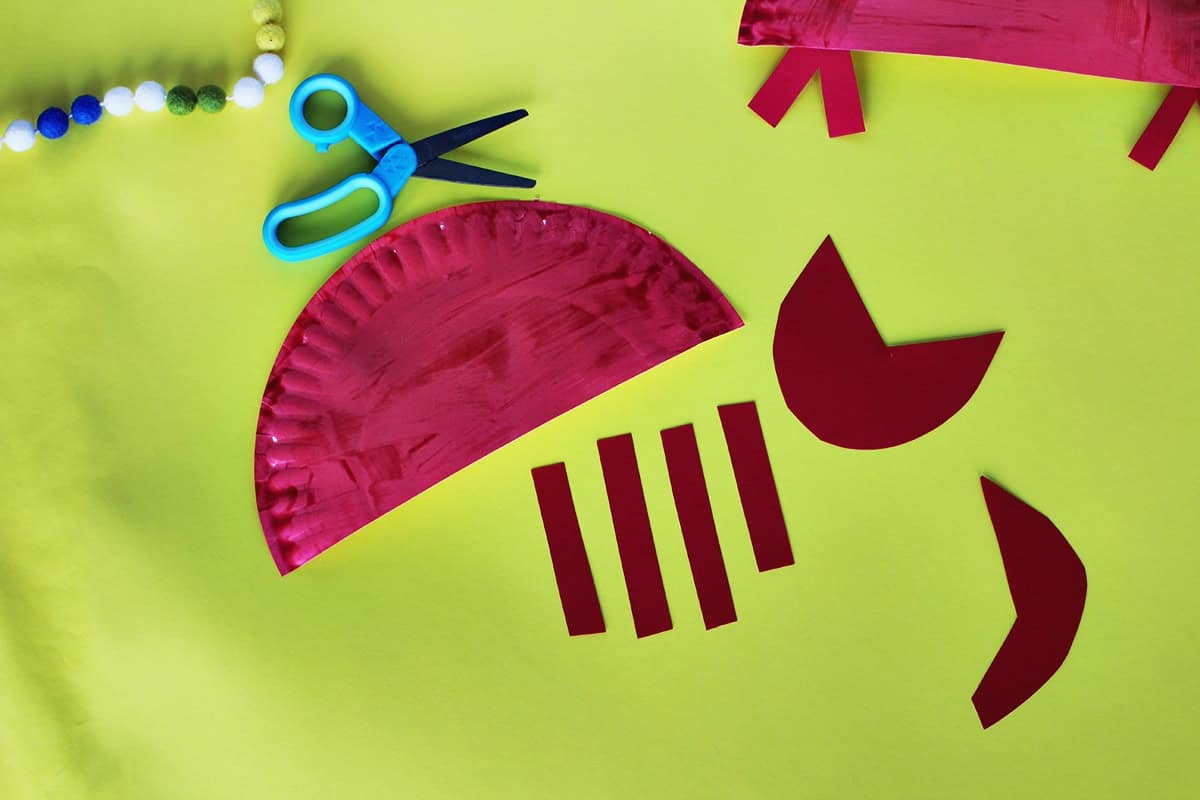 Paper plates painted red next to red cardstock paper to make craft crabs on a yellow background next to school glue and blue kids' scissors.