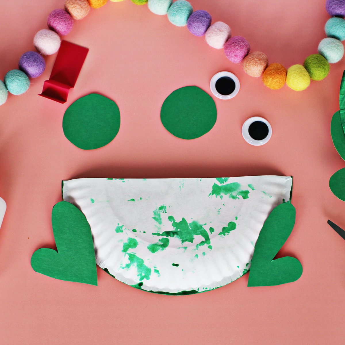 Back of a paper plate that has been cut in half with green paper glued onto it on a pink background.