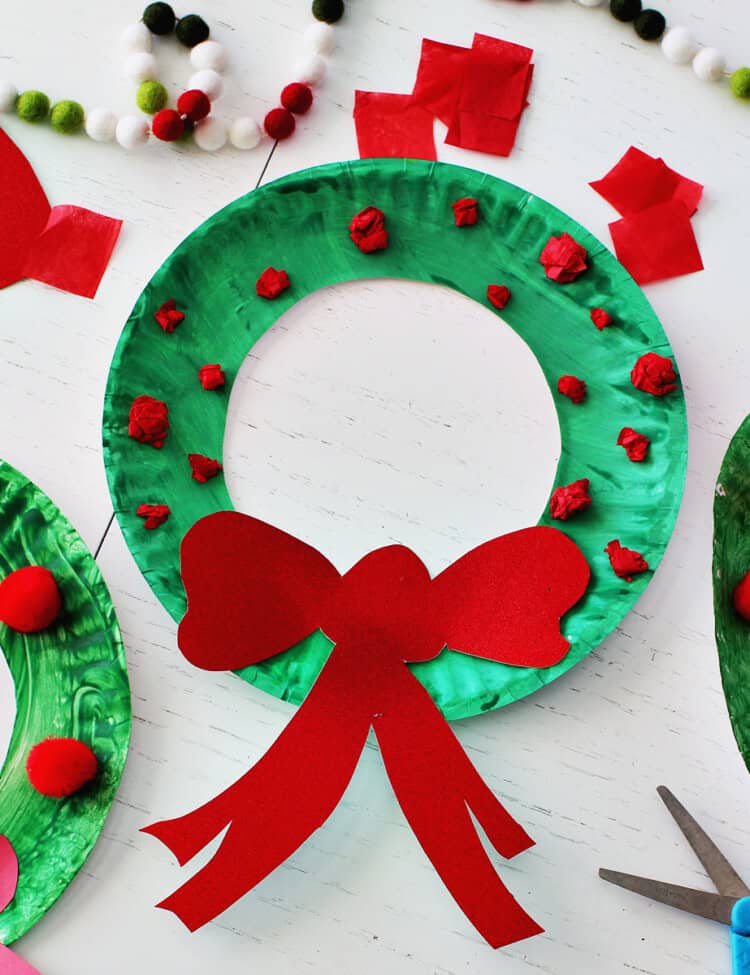 Green paper plate with center cut out and balled up red tissue paper along with a red cardstock bow glued onto it to make a wreath. Next to the paper plate is another green paper plate with red pom poms glued onto it, a bottle of school glue, red cardstock paper and red tissue paper squares.
