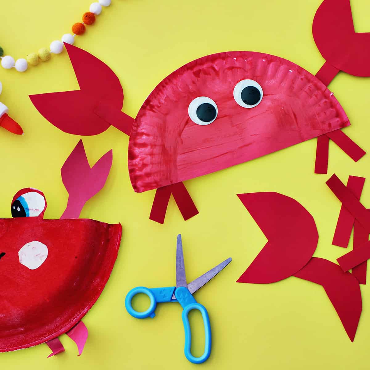 Paper plates painted red glued to red cardstock paper to make craft crabs on a yellow background next to school glue and blue kids' scissors.