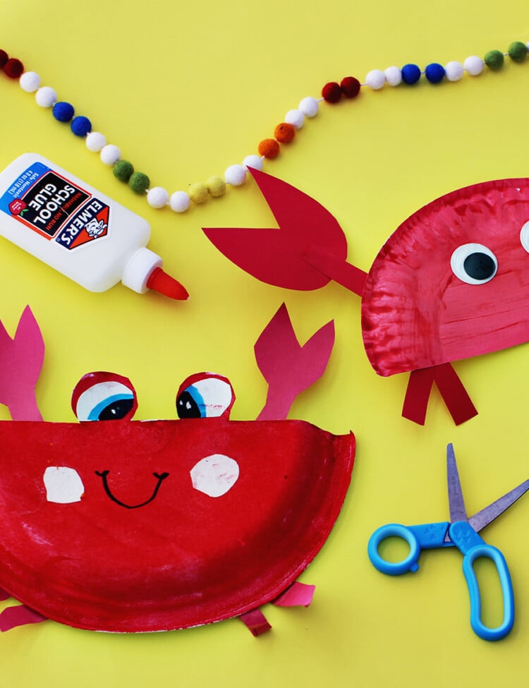 Paper plates painted red glued to red cardstock paper to make craft crabs on a yellow background next to school glue and blue kids' scissors.