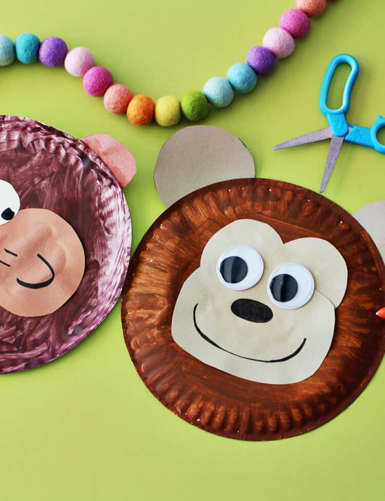 Two paper plates painted brown and decorated to look like monkeys next to each other and a pair of blue kids scissors on a light green background.