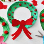 Green paper plate with center cut out and balled up red tissue paper along with a red cardstock bow glued onto it to make a wreath. Next to the paper plate is another green paper plate with red pom poms glued onto it, a bottle of school glue, red cardstock paper and red tissue paper squares.