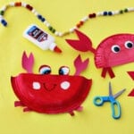 Paper plates painted red glued to red cardstock paper to make craft crabs on a yellow background next to school glue and blue kids' scissors.
