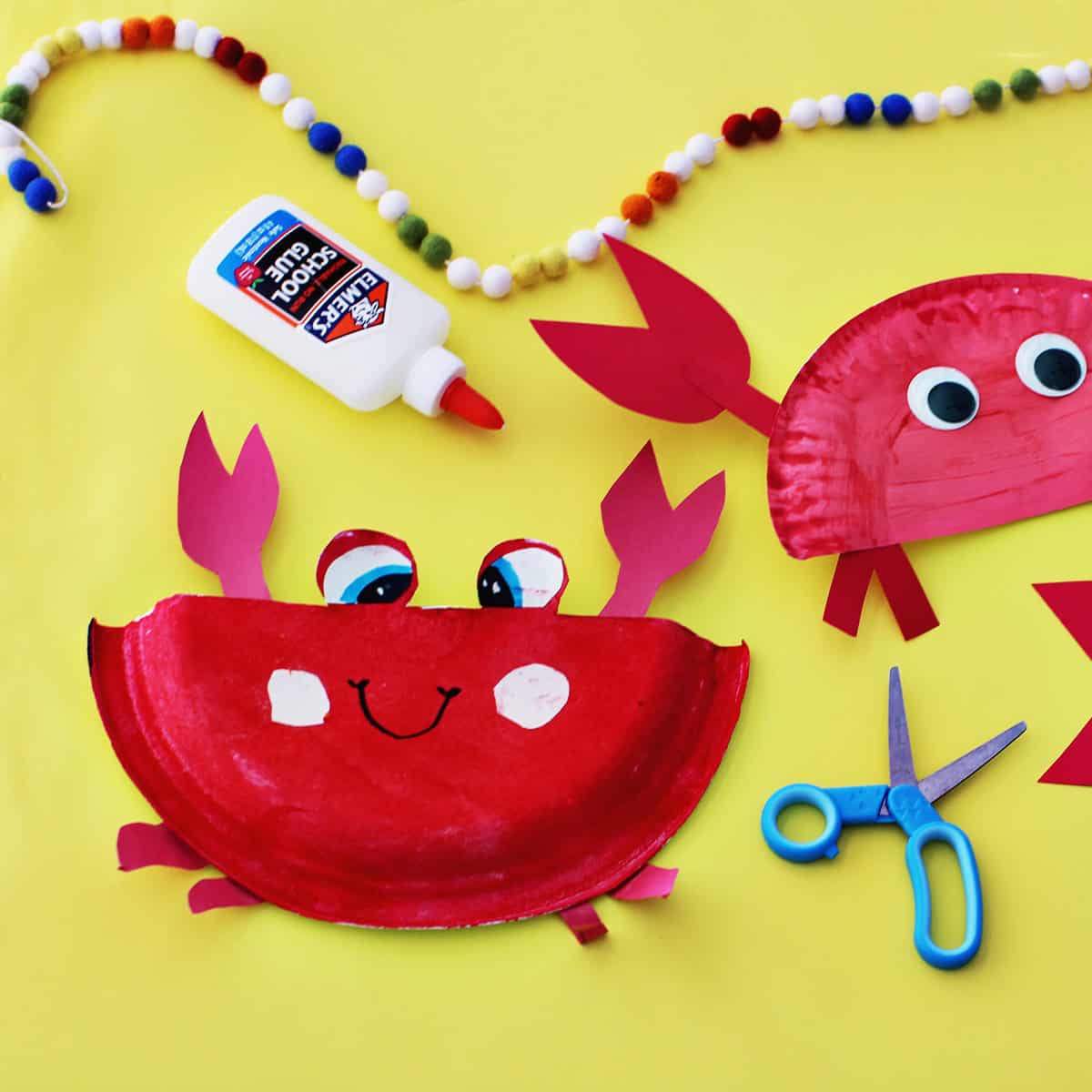 Paper plates painted red glued to red cardstock paper to make craft crabs on a yellow background next to school glue and blue kids' scissors.