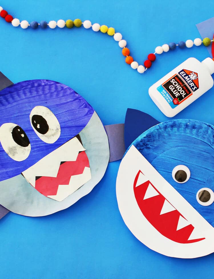 Blue painted paper plate with blue paper fins glued onto it and decorated to look like a shark. A bottle of school glue and another paper plate shark are next to the craft on a blue background.