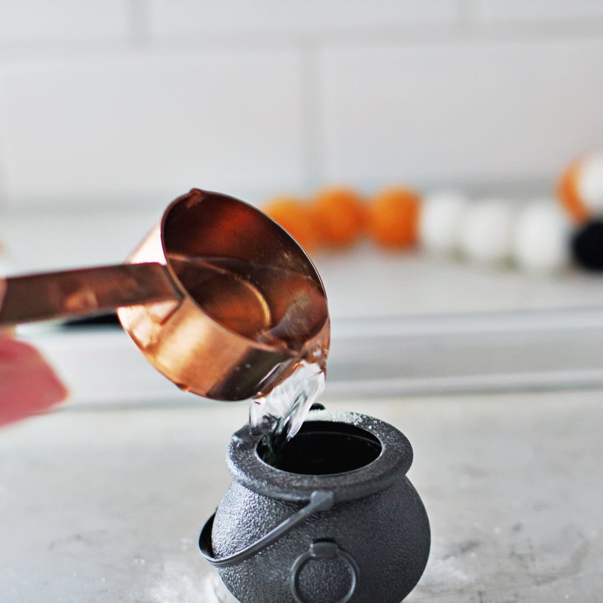 Vinegar being poured into a small black plastic cauldron sitting on a baking tray.