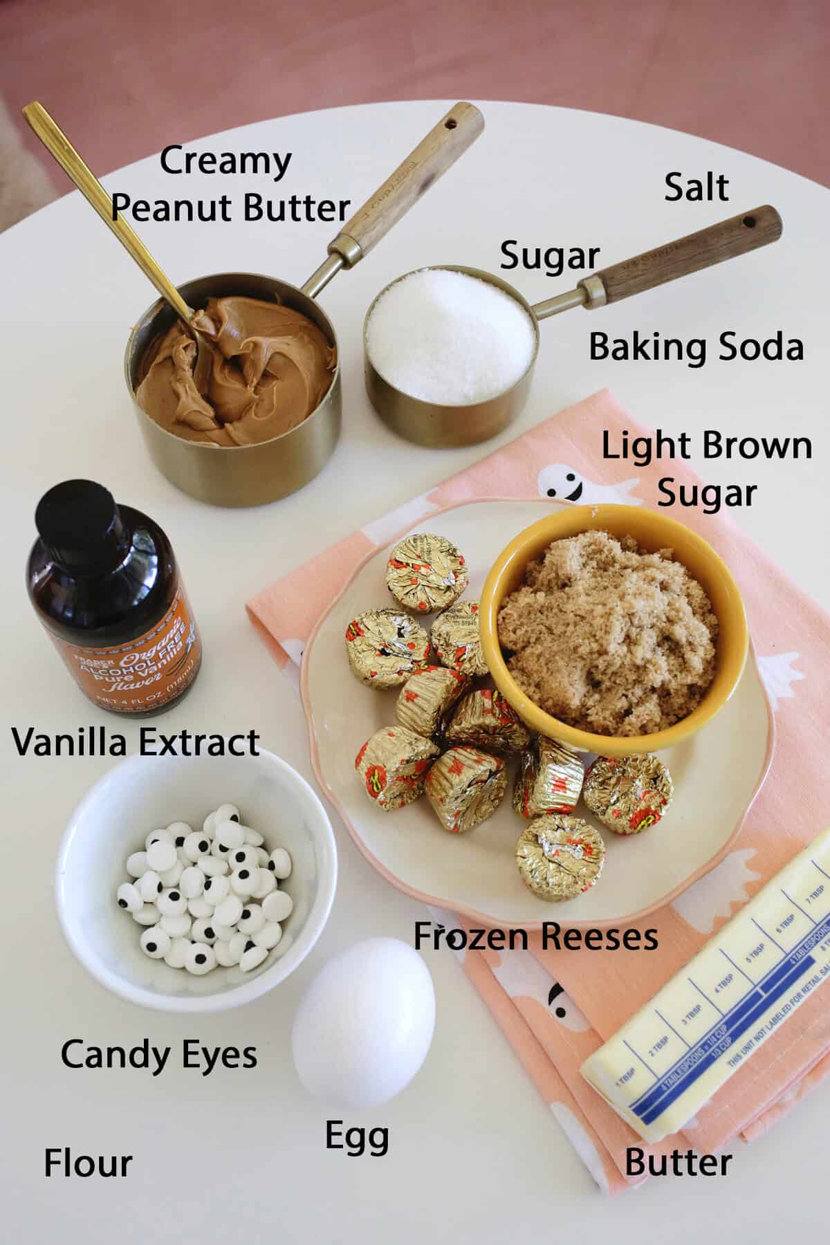peanut butter, sugar, salt, baking soda, light brown sugar, vanilla extract, candy eyes, reeses candies and a stick of butter on a table.