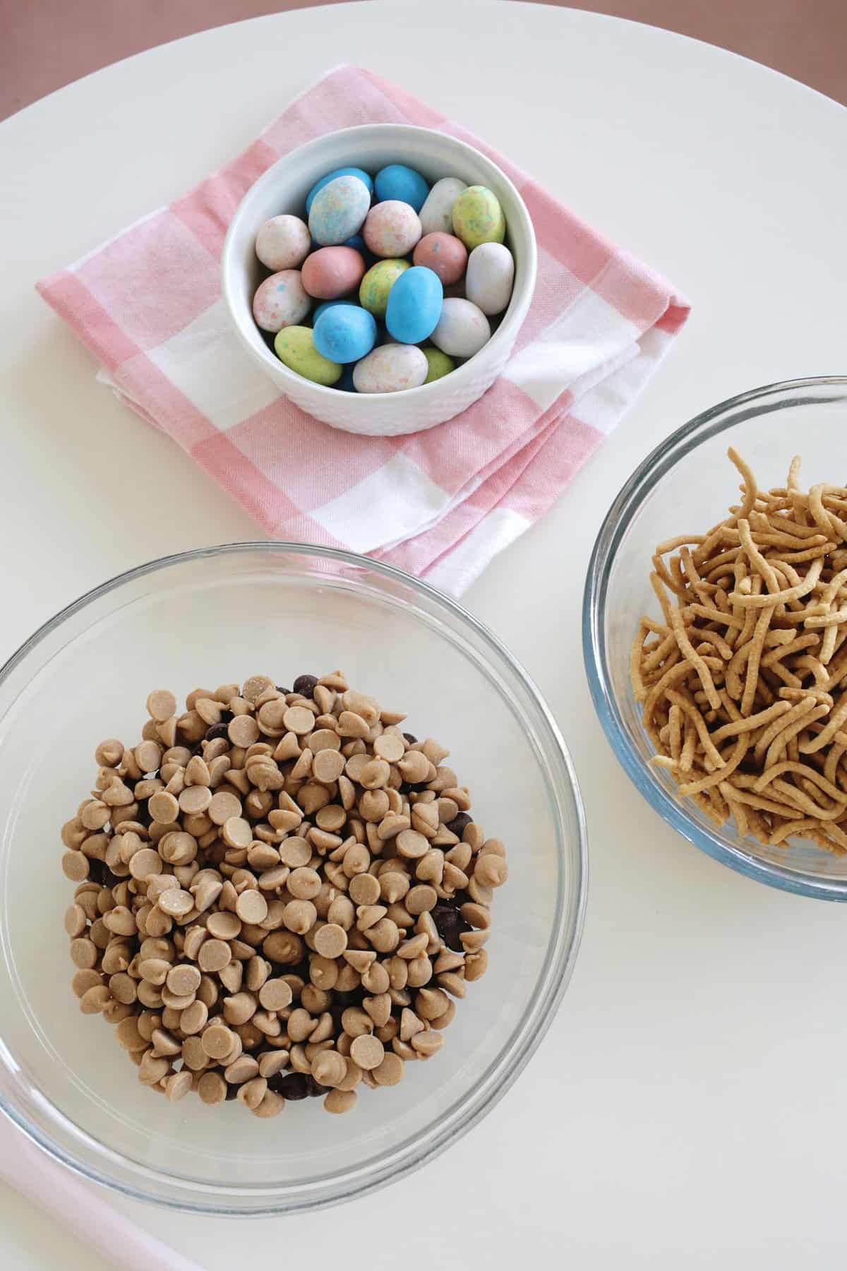 bowl of candy eggs next to a bowl of peanut butter and chocolate chips. Also sitting nearby is a bowl of chow mein noodles.