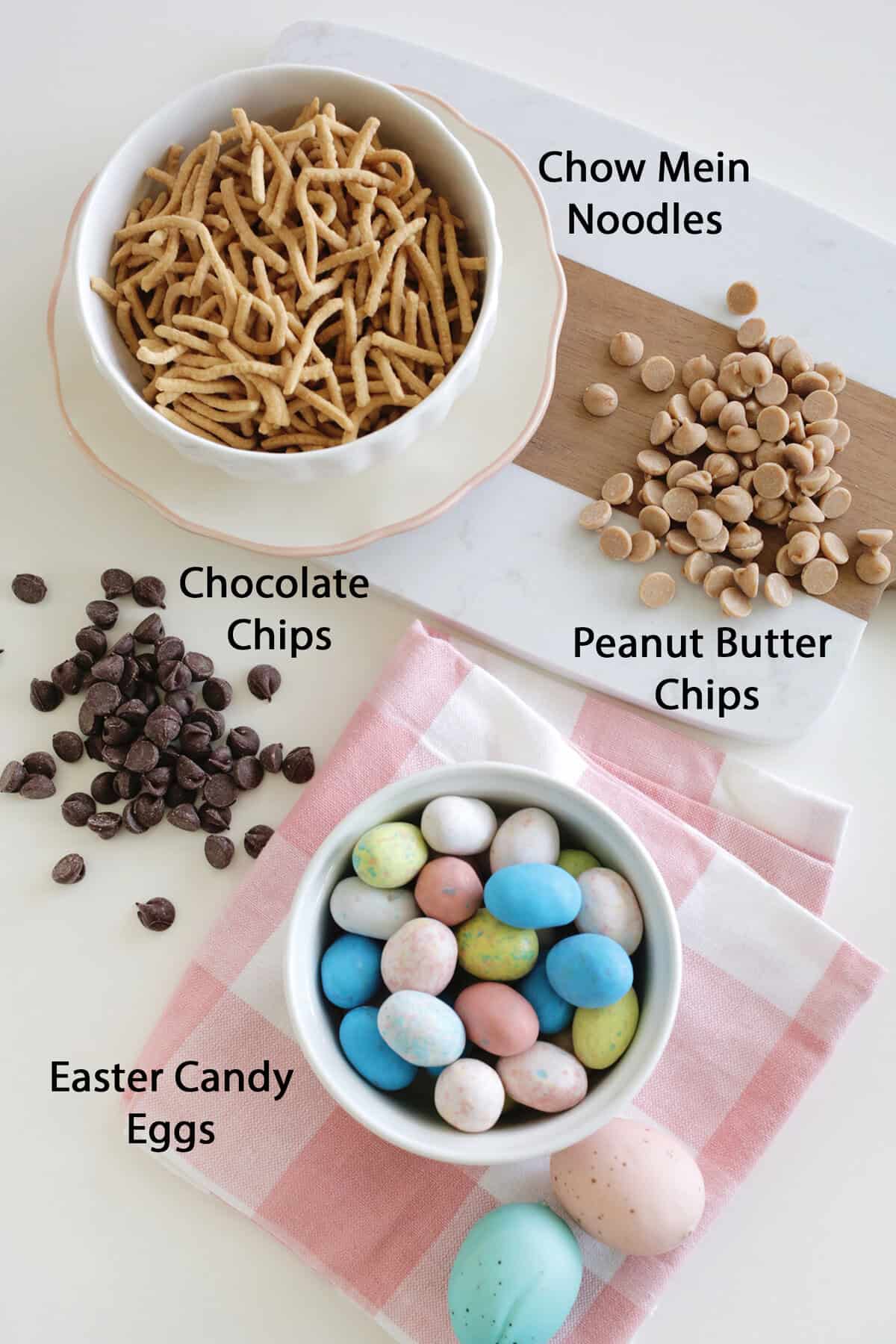 A bowl of chow mein noodles, a pile of peanut butter chips, chocolate chips and a small bowl of easter egg candies sitting on a table.