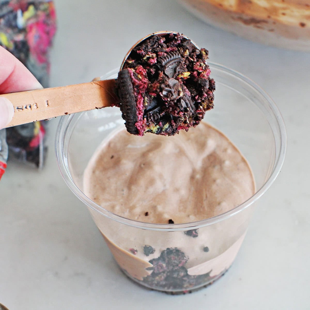 a tablespoon measuring cup adding crushed oreos to a clear cup holding pudding.